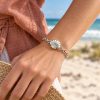 Silver-tone scallop shell chain bracelet worn on a woman's wrist beside a straw hat at the beach
