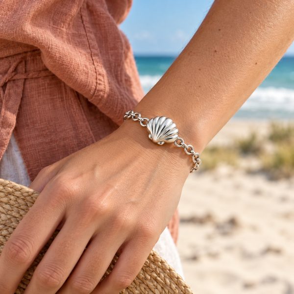 Silver-tone scallop shell chain bracelet worn on a woman's wrist beside a straw hat at the beach