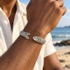 Silver-tone fish scale cuff bracelet worn on a man's wrist against a softly blurred beach background