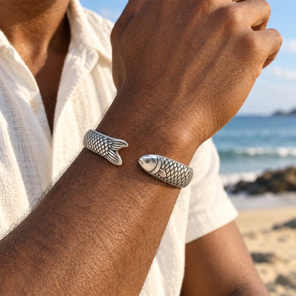 Silver-tone fish scale cuff bracelet worn on a man's wrist against a softly blurred beach background