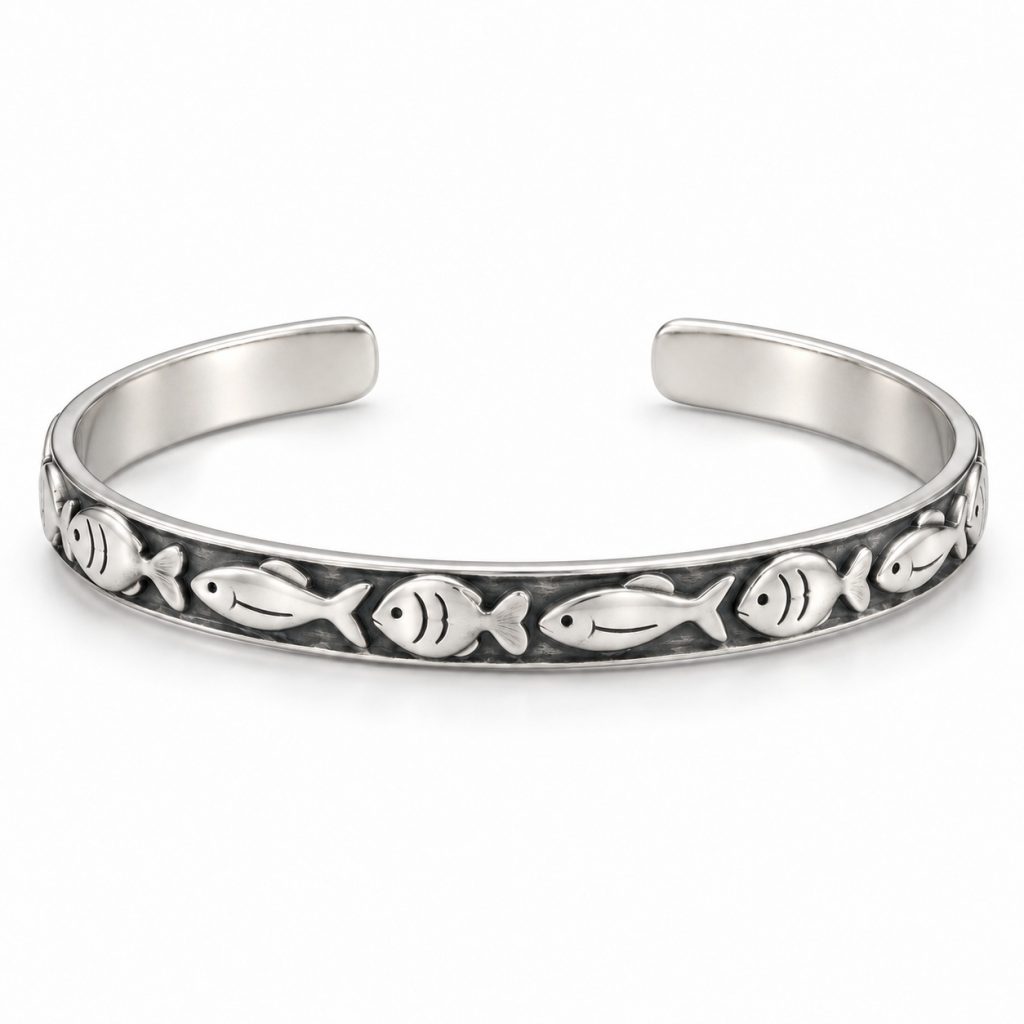 Silver-tone fish pattern cuff bracelet worn on a woman's wrist beside driftwood at the beach