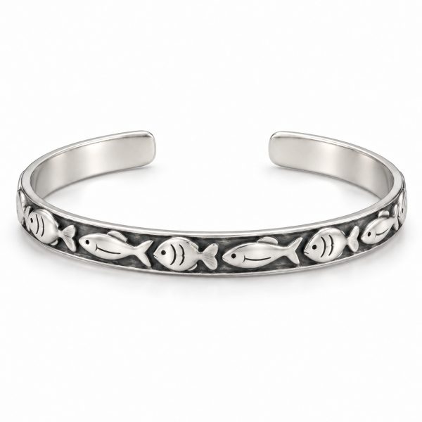 Silver-tone fish pattern cuff bracelet worn on a woman's wrist beside driftwood at the beach